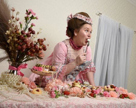 Close up portrait of cute female model wearing an opulent pink gown, the costume of a historical French baroque nobility. Eating indulgent food feast with pastries, flowers and rich jewels