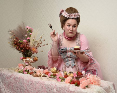 Nahaufnahme Porträt des niedlichen weiblichen Modells in einem opulenten rosa Kleid, das Kostüm eines historischen französischen Barockadels. Kuchen essen bei einem genussvollen Festmahl mit süßen Leckereien und reichhaltigen Speisen.