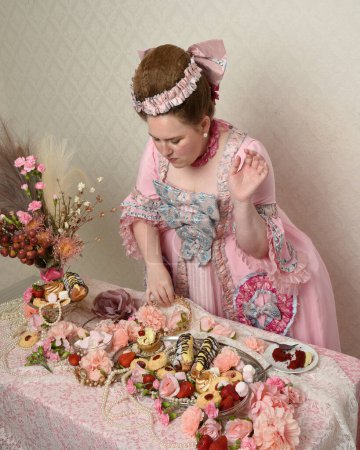 Primer plano retrato de modelo femenina linda con un vestido rosa opulento, el traje de una nobleza barroca francesa histórica. Comer pasteles en un banquete indulgente con dulces y alimentos ricos.
