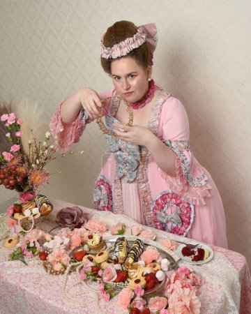 Nahaufnahme Porträt des niedlichen weiblichen Modells in einem opulenten rosa Kleid, das Kostüm eines historischen französischen Barockadels. Kuchen essen bei einem genussvollen Festmahl mit süßen Leckereien und reichhaltigen Speisen.