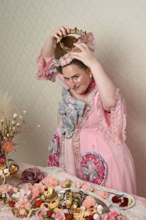Primer plano retrato de modelo femenina linda con un vestido rosa opulento, el traje de una nobleza barroca francesa histórica. Comer pasteles en un banquete indulgente con dulces y alimentos ricos.