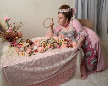 Primer plano retrato de modelo femenina linda con un vestido rosa opulento, el traje de una nobleza barroca francesa histórica. Comer pasteles en un banquete indulgente con dulces y alimentos ricos en mesa larga con papel pintado intrincado en el fondo.
