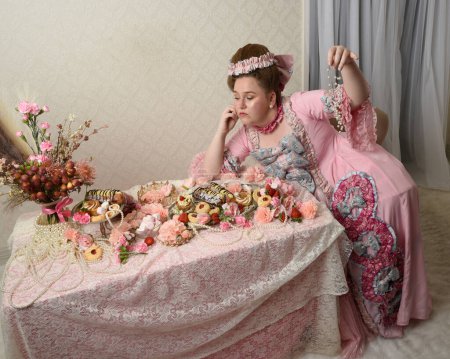 Primer plano retrato de modelo femenina linda con un vestido rosa opulento, el traje de una nobleza barroca francesa histórica. Comer pasteles en un banquete indulgente con dulces y alimentos ricos en mesa larga con papel pintado intrincado en el fondo.
