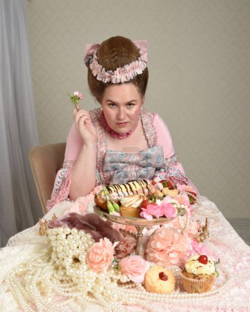 Close up portrait of cute female model wearing an opulent pink gown, the costume of a historical French baroque nobility.  sitting table with sweet cakes and indulgent food feast.