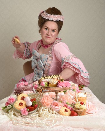 Close up portrait of cute female model wearing an opulent pink gown, the costume of a historical French baroque nobility.  sitting table with sweet cakes and indulgent food feast.