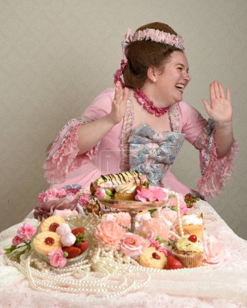 Gros plan portrait de mignonne mannequin féminine portant une robe rose opulente, le costume d'une noblesse baroque française historique. table assise avec des gâteaux sucrés et un festin de nourriture indulgent.