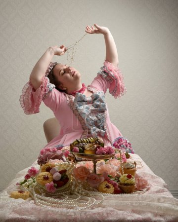 Primer plano retrato de modelo femenina linda con un vestido rosa opulento, el traje de una nobleza barroca francesa histórica. sentado a la mesa con pasteles dulces y comida indulgente.