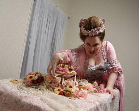 Nahaufnahme Porträt des niedlichen weiblichen Modells in einem opulenten rosa Kleid, das Kostüm eines historischen französischen Barockadels. bei süßen Kuchen und genussvollem Schlemmen am Tisch sitzen.