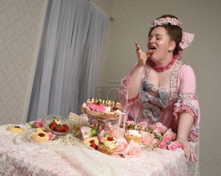 Gros plan portrait de mignonne mannequin féminine portant une robe rose opulente, le costume d'une noblesse baroque française historique. assis à table avec des gâteaux sucrés et un festin de nourriture indulgent.