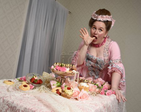 Nahaufnahme Porträt des niedlichen weiblichen Modells in einem opulenten rosa Kleid, das Kostüm eines historischen französischen Barockadels. bei süßen Kuchen und genussvollem Schlemmen am Tisch sitzen.
