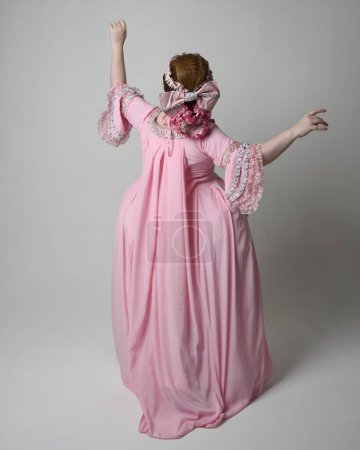 Photo for Full length portrait of woman wearing historical French baroque pink gown in style of Marie Antoinette with elegant hairstyle. Standing pose, walking away from the camera, isolated on studio background. - Royalty Free Image