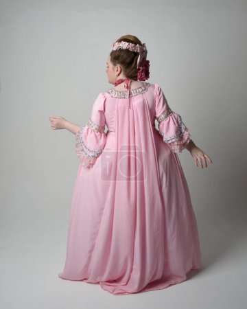 Photo for Full length portrait of woman wearing historical French baroque pink gown in style of Marie Antoinette with elegant hairstyle. Standing pose, walking away from the camera, isolated on studio background. - Royalty Free Image
