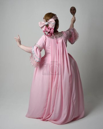 Ganzkörperporträt einer Frau, die ein historisches französisches barockes rosafarbenes Kleid im Stil von Marie Antoinette mit eleganter Frisur trägt. Stehende Pose, weg von der Kamera, isoliert auf Studiohintergrund.