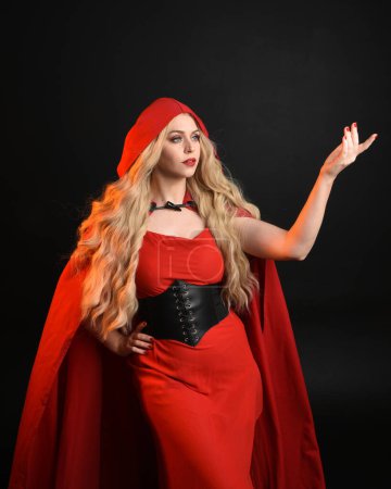 close up portrait of beautiful model  with long blonde hair wearing glamorous fantasy dress with corset and  flowing red riding hood fairytale cloak. isolated on dark studio background.