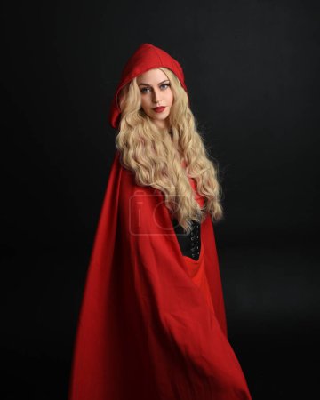 Nahaufnahme Porträt der schönen Modell mit langen blonden Haaren trägt glamouröse Fantasy-Kleid mit Korsett und fließenden Rotkäppchen märchenhaften Mantel. isoliert auf dunklem Studiohintergrund.
