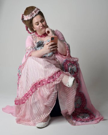 Full length portrait female model wearing opulent pink gown costume of historical French baroque nobility, style of Marie Antoinette. Sitting pose on throne using modern mobile phone technology.