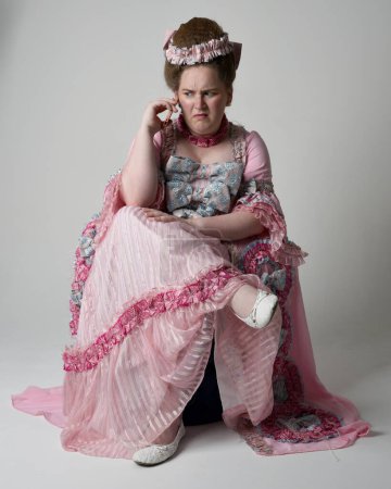 Retrato de cuerpo entero modelo femenino con opulento traje de bata rosa de la nobleza barroca francesa histórica, estilo de María Antonieta. Posar sentado en el trono utilizando la tecnología moderna de telefonía móvil.