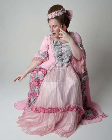 Retrato de cuerpo entero modelo femenino con opulento traje de bata rosa de la nobleza barroca francesa histórica, estilo de María Antonieta. Posar sentado en el trono utilizando la tecnología moderna de telefonía móvil.