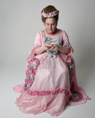 Full length portrait female model wearing opulent pink gown costume of historical French baroque nobility, style of Marie Antoinette. Sitting pose on throne using modern mobile phone technology.