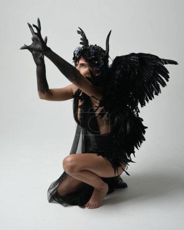 Photo for Full length portrait of female model wearing gothic horned headdress with halloween black dress and fantasy angel feather wings. Crouching pose, kneeling on floor. Isolated studio background - Royalty Free Image