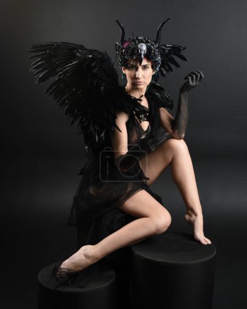Photo for Full length portrait of model wearing gothic horned headdress with halloween black dress, fantasy angel feather wings. Crouching pose. Isolated dark studio background, cinematic shadow silhouettes - Royalty Free Image