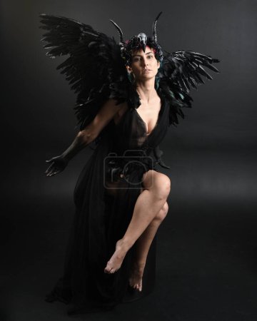 Photo for Full length portrait of model wearing gothic horned headdress with halloween black dress, fantasy angel feather wings. Crouching pose. Isolated dark studio background, cinematic shadow silhouettes - Royalty Free Image