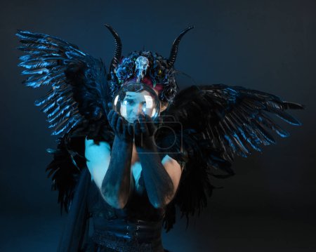 Photo for Close up portrait of female model wearing gothic horned headdress, halloween black dress, fantasy angel feather wings. Holding mystical crystal ball. dark studio background, cinematic shadow silhouette - Royalty Free Image