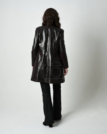 Photo for Full length portrait of brunette woman wearing long leather trench coat and black boots. Standing pose walking away from camera, facing backwards. Isolated silhouette on white studio background. - Royalty Free Image