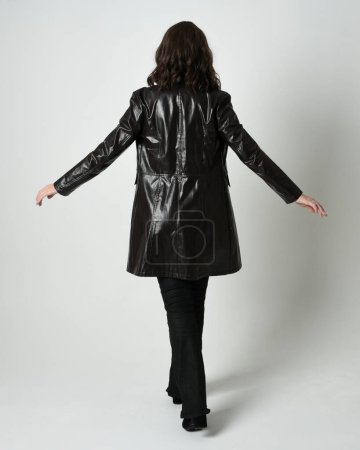 Photo for Full length portrait of brunette woman wearing long leather trench coat and black boots. Standing pose walking away from camera, facing backwards. Isolated silhouette on white studio background. - Royalty Free Image