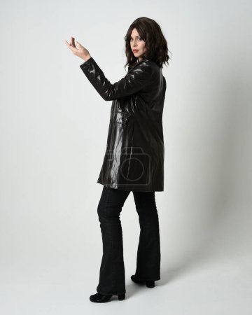 Photo for Full length portrait of brunette woman wearing long leather trench coat. Standing pose walking away from camera, looking back over shoulder, reaching out. Isolated on white studio background. - Royalty Free Image