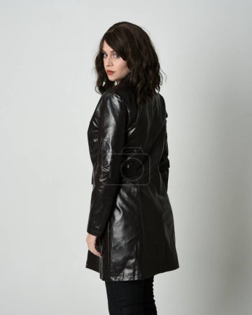 Photo for Close up portrait of brunette woman wearing long leather trench coat. Standing pose in side profile, looking back over shoulder, reaching out. Isolated on white studio background. - Royalty Free Image