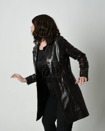 Photo for Close up portrait of brunette woman wearing long leather trench coat. Standing pose in side profile, looking back over shoulder, reaching out. Isolated on white studio background. - Royalty Free Image