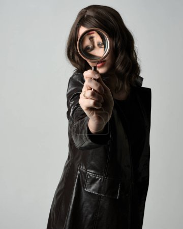 Nahaufnahme Porträt der schönen brünetten Frau in schwarzem Leder Trenchcoat. Mit detektivischer Lupe, ausgreifend in Richtung Kamera, auf der Suche nach Entdeckung. Isolierter Studiohintergrund
