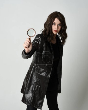 close up portrait of beautiful brunette woman wearing black leather trench coat. Holding detective magnifying glass, reaching out towards camera, searching for discovery. Isolated  studio background