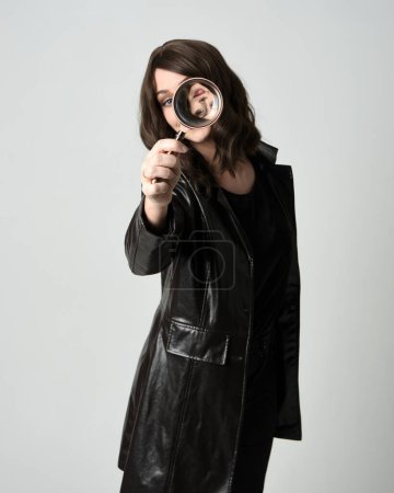 Nahaufnahme Porträt der schönen brünetten Frau in schwarzem Leder Trenchcoat. Mit detektivischer Lupe, ausgreifend in Richtung Kamera, auf der Suche nach Entdeckung. Isolierter Studiohintergrund