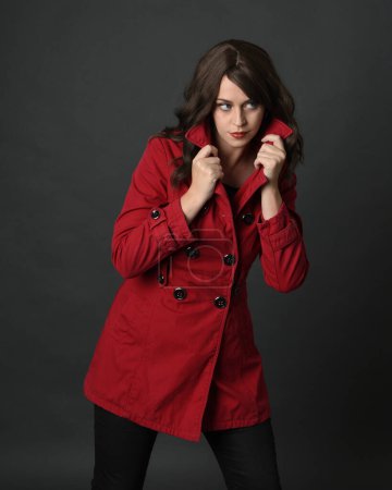 close up portrait of beautiful brunette woman model, wearing red trench coat jacket.isolated on dark studio background with shadows.  holding collar in secretive hiding pose.