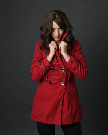 close up portrait of beautiful brunette woman model, wearing red trench coat jacket.isolated on dark studio background with shadows.  holding collar in secretive hiding pose.