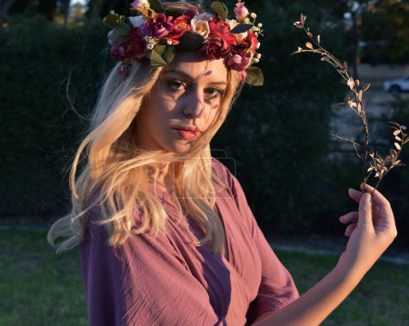 Téléchargez les photos : Portrait rapproché d'une jolie mannequin femme blonde portant une couronne de fleurs et une robe violette. des plantes et des arbres verts en arrière-plan - en image libre de droit
