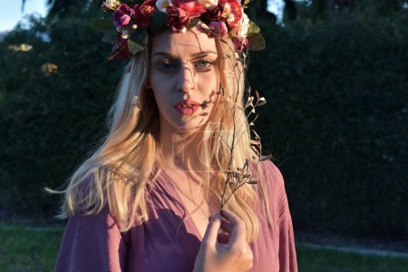close up portrait of pretty blonde female model wearing a flower crown wreath and purple dress.  green nature  plants and trees in background