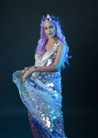Photo for Artistic portrait of beautiful female model with long purple hair wearing a fantasy fairy crown, wearing a rainbow glitter sequin ball gown. gestural flowing pose, isolated on dark studio background. - Royalty Free Image