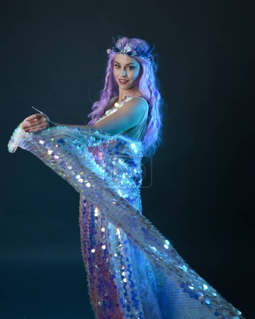 artistic portrait of beautiful female model with long purple hair wearing a fantasy fairy crown, wearing a rainbow glitter sequin ball gown. gestural flowing pose, isolated on dark studio background.