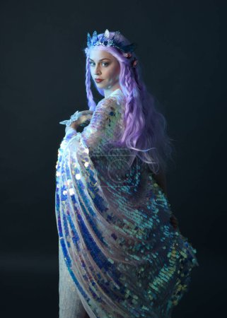 artistic portrait of beautiful female model with long purple hair wearing a fantasy fairy crown, wearing a rainbow glitter sequin ball gown. gestural flowing pose, isolated on dark studio background.