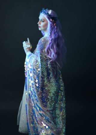 retrato artístico de la hermosa modelo femenina con el pelo largo de color púrpura con una corona de hadas de fantasía, con un vestido de lentejuelas arco iris brillo. pose gestual fluida, aislada sobre fondo oscuro del estudio.