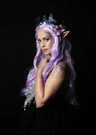 Photo for Artistic portrait of beautiful female model with long purple hair wearing a fantasy fairy crown and elf ears,  gestural  pose with hands reaching up to touch face.  isolated on dark studio background. - Royalty Free Image