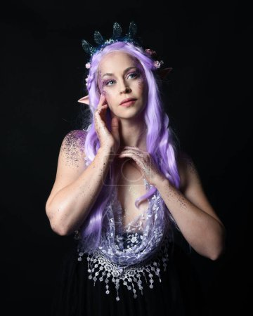 Photo for Artistic portrait of beautiful female model with long purple hair wearing a fantasy fairy crown and elf ears,  gestural  pose with hands reaching up to touch face.  isolated on dark studio background. - Royalty Free Image