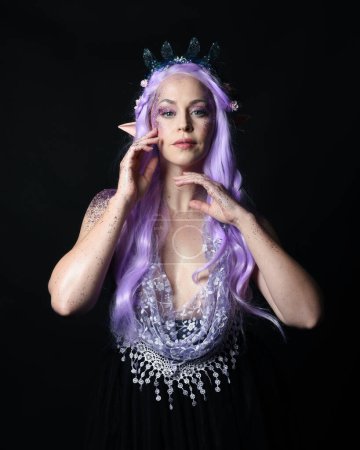 portrait artistique d'une belle mannequin féminine aux longs cheveux violets portant une couronne de fée fantaisie et des oreilles d'elfe, pose gestuelle avec les mains allant jusqu'au visage tactile. isolé sur fond sombre studio.