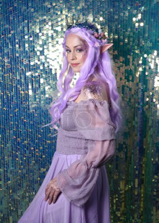  Porträt des süßen weiblichen Modells mit langen lila Haaren, das eine fantasievolle Blumenkrone mit Elfenohren trägt. Isoliert auf funkelndem Regenbogen-Pailletten-Hintergrund mit Glitzern.