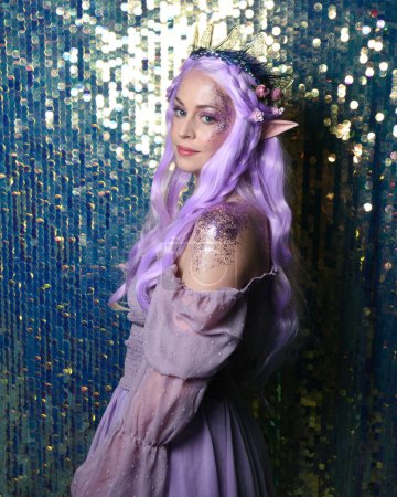 Photo for Portrait of cute female model with long purple hair wearing a fantasy fairy flower crown with elf ears. Isolated on sparkling rainbow sequin background with glitter. - Royalty Free Image
