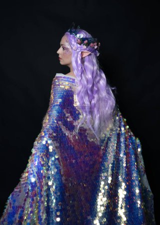 Photo for Artistic portrait of beautiful female model with long purple hair and elf ears, wearing a fantasy fairy crown, wearing a rainbow glitter sequin ball gown. Standing in side profile, isolated on dark studio background. - Royalty Free Image