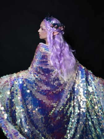 Photo for Artistic portrait of beautiful female model with long purple hair and elf ears, wearing a fantasy fairy crown, wearing a rainbow glitter sequin ball gown. Standing in side profile, isolated on dark studio background. - Royalty Free Image
